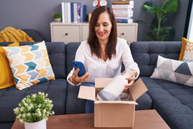 Genç, güzel, İspanyol bir kadın akıllı telefon kullanarak evdeki karton kutuları açıyor.