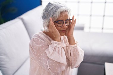 Middle age grey-haired woman suffering for headache sitting on sofa at home