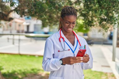 Parkta akıllı telefon kullanarak gülümseyen Afrikalı Amerikalı kadın doktor.