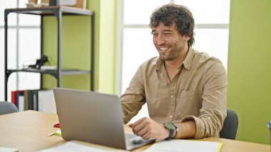 Young hispanic man business worker smiling confident sitting on table at office