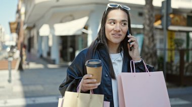 Genç, güzel, İspanyol bir kadın elinde alışveriş poşetleri ve kahveyle akıllı telefondan konuşuyor.