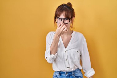 Young beautiful woman wearing casual shirt over yellow background smelling something stinky and disgusting, intolerable smell, holding breath with fingers on nose. bad smell  clipart