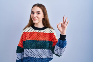 Mavi arka planda duran Latin asıllı genç kız gülümsüyor ve el ve parmaklarıyla 