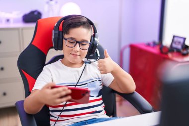Genç İspanyol çocuk akıllı telefonuyla video oyunu oynuyor mutlu ve olumlu gülümsüyor, başparmağıyla mükemmel ve onay işareti yapıyor. 