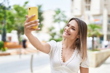 Beyaz kadın gülümsüyor. Sokakta akıllı telefonun yanında özçekim yapıyor.