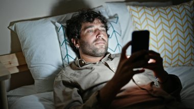 Genç İspanyol adam yatakta akıllı telefon kullanıyor.
