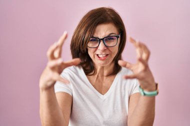 Orta yaşlı İspanyol kadın pembe arka planda duruyor öfkeden çıldırmış, elleri boğazlamaya çalışıyor, deli gibi bağırıyor. 