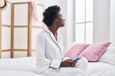 Young african american woman using smartphone sitting on bed at bedroom