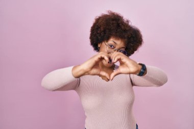 Young african american woman standing over pink background smiling in love doing heart symbol shape with hands. romantic concept. 
