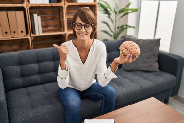 Terapi ofisinde çalışan esmer bir kadın beyni tutup baş parmağını yukarı kaldırıyor ve ağzı açık gülümsüyor. 