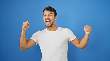 Genç İspanyol adam gülümsüyor. Kendine güveni tam. Yalıtılmış mavi arka planda bir jest yapıyor.