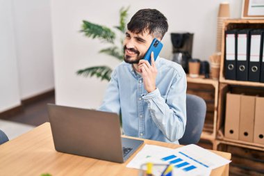 Genç İspanyol iş adamı ofisteki bilgisayarla akıllı telefondan konuşuyor.