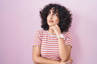 Orta Doğulu genç bir kadın pembe arka planda, elleri çenesinde soru sorarken, düşünceli bir ifade takınıyor. Düşünceli bir yüzle gülümsüyor. şüphe kavramı. 