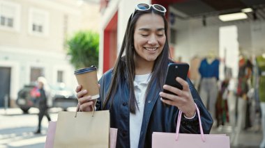 Genç ve güzel İspanyol kadın akıllı telefon kullanıyor. Elinde alışveriş poşetleri ve sokakta kahve tutuyor.