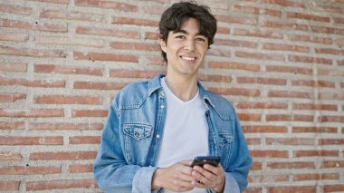 Zeki telefon kullanan genç bir İspanyol, izole tuğla arka planda gülümsüyor.