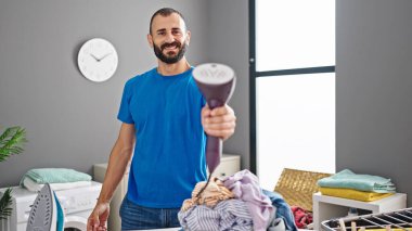 Çamaşır odasında gülümseyen, dikey ütü makinesi tutan İspanyol genç adam.