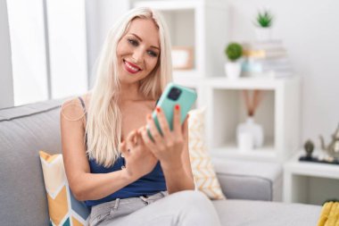 Evdeki kanepede oturan akıllı telefon kullanan genç sarışın kadın.