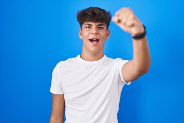 Mavi arka planda öfkeli ve kızgın duran İspanyol genç öfkeyle bağırırken yumruğunu sallıyor ve öfkeleniyor. öfke ve agresif konsept. 