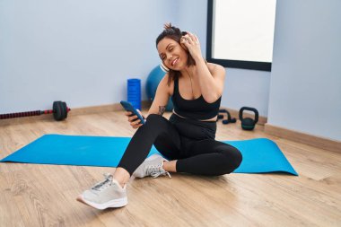 Young beautiful hispanic woman sitting on yoga mat listening to music at sport center