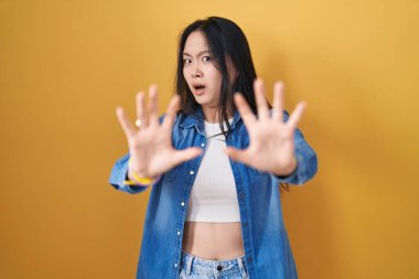 Sarı arka planda duran genç Asyalı kadın korku ifadesinden korkuyor ve dehşete düşmüş. El hareketlerini durdur, şok içinde bağır. panik konsepti. 