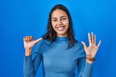 Genç Brezilyalı bir kadın mavi arka planda duruyor. Altıncı parmaklarını gösterip işaret ederken kendinden emin ve mutlu gülümsüyor.. 