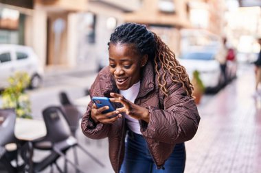 Afro-Amerikalı kadın sokakta neşeli bir ifadeyle akıllı telefon kullanıyor.