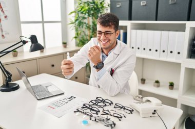 Young hispanic man optician smiling confident holding eye drops at clinic