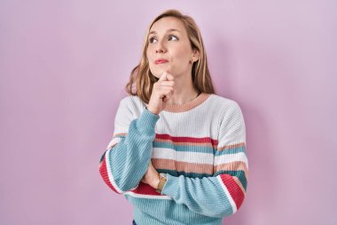 Pembe arka planda duran genç sarışın kadın, elleri çenesinde soru sorarken, düşünceli bir yüz ifadesi vardı. Gülümseyen ve düşünceli bir yüz. şüphe kavramı. 