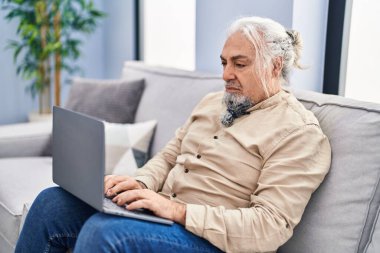 Evdeki koltukta oturan orta yaşlı, kır saçlı adam dizüstü bilgisayar kullanıyor.