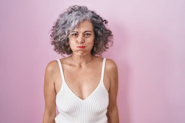 Pembe arka planda duran gri saçlı, komik yüzlü orta yaşlı bir kadın. Havayla şişmiş ağız, çılgın bir ifade.. 