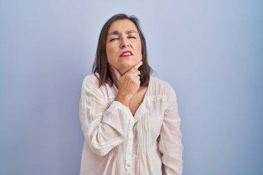 Middle age hispanic woman standing over blue background touching painful neck, sore throat for flu, clod and infection 