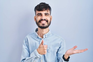 Mavi arka planda duran sakallı genç İspanyol adam avuç içini gösteriyor ve baş parmaklarıyla iyi bir hareket yapıyor, mutlu ve neşeli gülümsüyor. 
