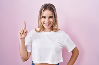 Pembe arka planda duran genç sarışın kadın bir numarayı gösterip işaret ederken kendinden emin ve mutlu bir şekilde gülümsüyor.. 