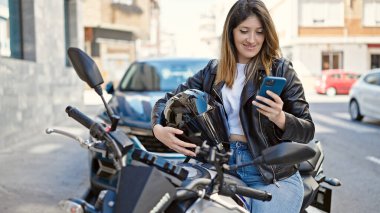 Sokakta motosiklette oturan akıllı telefon kullanan genç sarışın kadın.