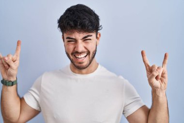 Hispanic man with beard standing over white background shouting with crazy expression doing rock symbol with hands up. music star. heavy music concept. 