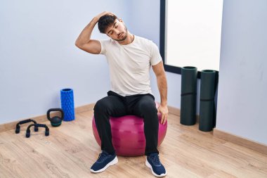 Young hispanic man sitting on fit ball stretching at sport center