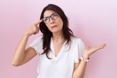 Orta yaşlı İspanyol kadın, günlük beyaz tişört ve gözlük takıyor. Kafası karışmış ve avuç içi açık bir şekilde fotokopi alanını gösteriyor ve alnını işaret ediyor. Bunu bir düşün.. 