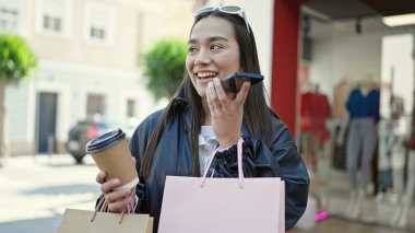 Güzel İspanyol bir kadın elinde alışveriş poşetleri ve sokakta kahve tutarak akıllı telefonuyla sesli mesaj yolluyor.