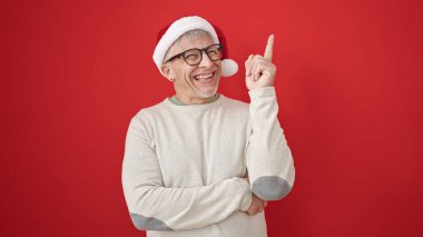 Orta yaşlı, gri saçlı, yılbaşı şapkası takan, izole edilmiş kırmızı arka plan üzerinde fikir ifadesi olan bir adam.