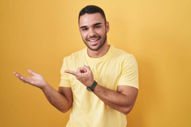 Young hispanic man standing over yellow background amazed and smiling to the camera while presenting with hand and pointing with finger. 