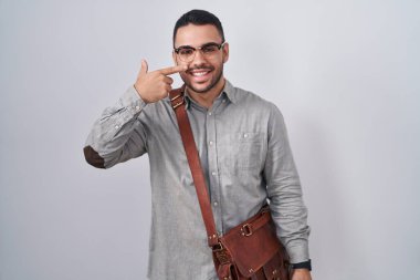 İspanyol asıllı genç bir adam bavul takıyor, parmaklarını burnuna ve yüzüne gösteriyor, neşeli bir şekilde gülümsüyor. güzellik konsepti 