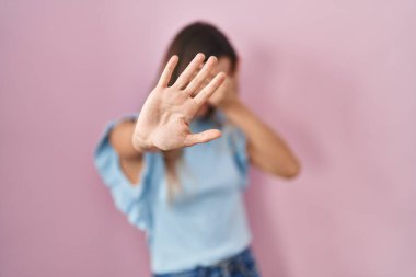 Pembe arka planda duran genç İspanyol kadın elleriyle gözlerini kapatıyor ve üzgün ve korku ifadesiyle dur hareketi yapıyor. utangaç ve olumsuz kavram. 