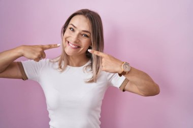 Pembe arka planda duran, neşeli gülümseyen ve parmaklarıyla ve ağzıyla işaret eden sarışın beyaz kadın. diş sağlığı kavramı. 