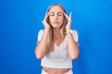 Young caucasian woman standing over blue background with hand on head, headache because stress. suffering migraine. 
