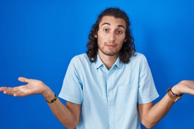 Young hispanic man standing over blue background clueless and confused expression with arms and hands raised. doubt concept. 