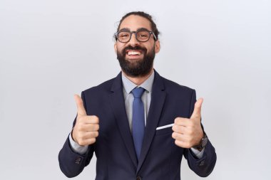 Hispanic man with beard wearing suit and tie success sign doing positive gesture with hand, thumbs up smiling and happy. cheerful expression and winner gesture. 