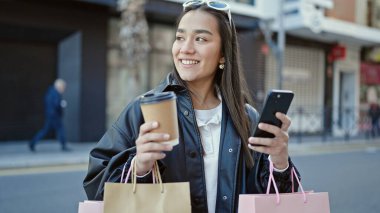 Genç ve güzel İspanyol kadın akıllı telefon kullanıyor. Elinde alışveriş poşetleri ve sokakta kahve tutuyor.