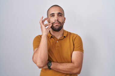 Hispanic man with beard standing over white background mouth and lips shut as zip with fingers. secret and silent, taboo talking 