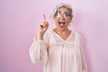 Pembe arka planda duran gri saçlı orta yaşlı kadın başarılı bir fikri işaret ediyor. Heyecanlı ve mutlu. Bir numara.. 