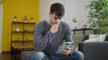Genç İspanyol adam akıllı telefon kullanıyor. Evdeki kanepede oturup endişeleniyor.